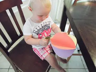 A little girl with blonde hair, a white t-shirt, and pink shirt sits in a chair, holding a paper hot air balloon craft made with orange, purple, and yellow paper.