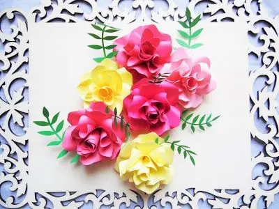 A light wooden board with ornate sides holds a bundle of paper Ansley roses and green paper leaves in the center. Paper flowers like these are colorful and appear delicate but sturdy. 