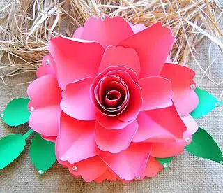 A closeup of a blooming dark pink scarlet rose made of paper. Paper leaves are under the flower, and straw is nestled above the paper rose. 