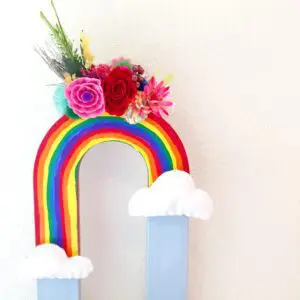This cardboard letter vase is in the shape of a “U,” turned upside-down and painted to look like a rainbow in the sky with clouds. A colorful bouquet of paper flowers sits at the very top of the rainbow.