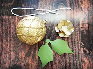 A round gold latticed ornament surrounded by a piece of string, a gold flower, and green paper leaves.