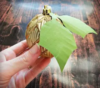 Abbi Kirsten's hand is holding the finished gold ornament with attached paper leaves. The ornament is jazzed up with paper flowers and greenery.