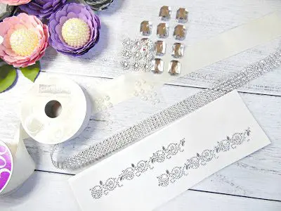 A view of some of the supplies needed from above. The items include paper flowers, a rhinestone ribbon, ribbon spools, and rhinestone sticker designs.