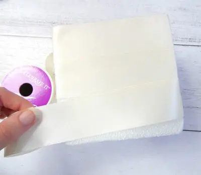 Abbi's fingers are seen carefully wrapping a floral block with wide satin ribbon while it rests on a white table. This is the base for the DIY wedding centerpiece craft.
