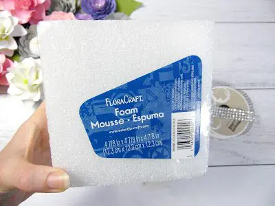 Next to a bundle of faux paper flowers, Abbi Kirsten's hand holds a styrofoam Floracraft cube used in the DIY wedding centerpiece.