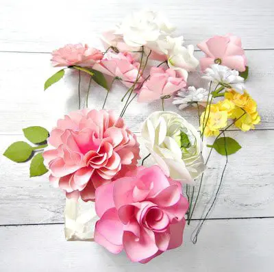 Dahlia, Ansley rose, ranunculus, and hydrangea stemmed paper flowers, made using Abbi Kirsten's templates, lay on a white wooden table.