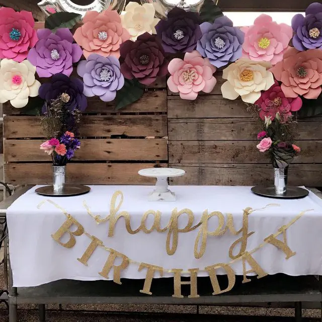 A table covered in a white tablecloth is decorated with a gold "happy birthday" banner and flower arrangements. On the wooden wall behind a table, a giant paper flower banner hangs, made up of colorful paper flowers.