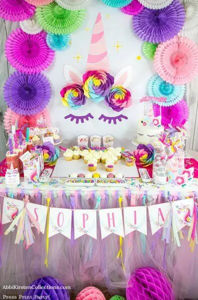 A fancy party wall featuring many paper crafts like rainbow paper flowers, cupcakes, a unicorn cake, and whimsical unicorn themed decorations. A paper sign saying "Sophia" hangs on the table.
