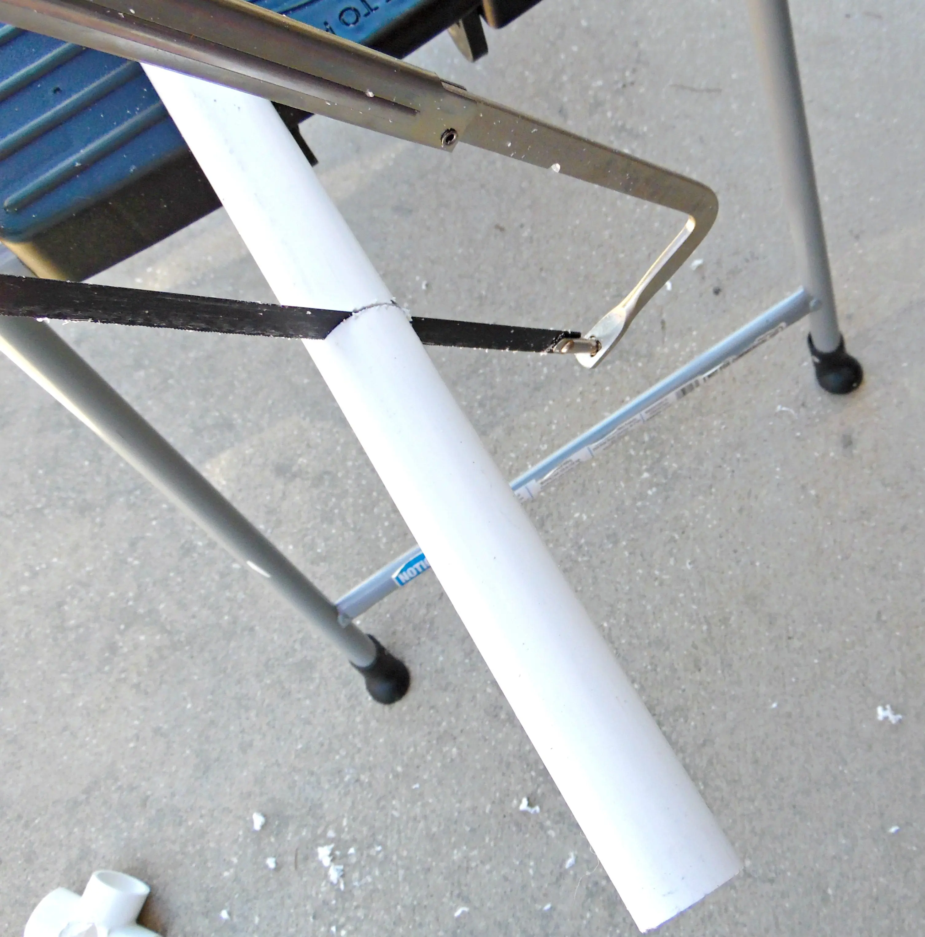 A piece of white PVC is placed on the end of a workbench while a handsaw cuts off an 8 to 9-inch piece. This is how to make the legs of the giant paper flower stem display stand.