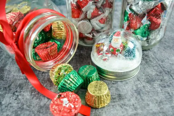 A closeup of a snowglobe lid and mason jars filled with red, green, and gold glimmering candies and red ribbons. These DIY snowglobe mason jar crafts are perfect gifts.  