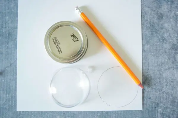 A piece of white cardstock lays on a gray tabletop. On the paper is a pencil, a mason jar lid, and half of a clear fillable ornament. 