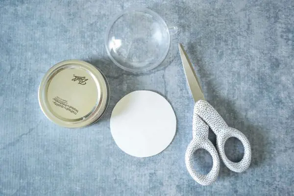 These are some supplies needed for a DIY snowglobe mason jar craft. A jarl lid, scissors, a cut cardstock circle, and a fillable clear ornament half are on a gray table. 