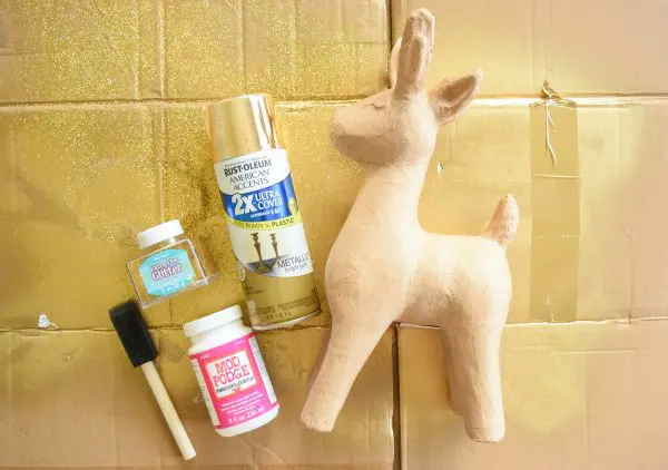 A paper mache deer laying on a cardboard piece with gold spray paint and other supplies. 
