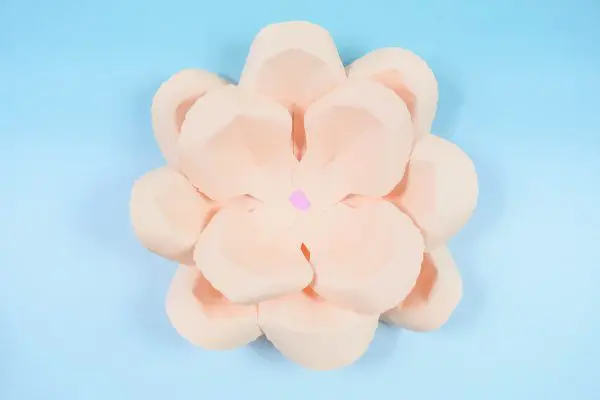 Two layers of a peach DIY paper rose serena flower is seen from above, sitting on a light blue tabletop. 