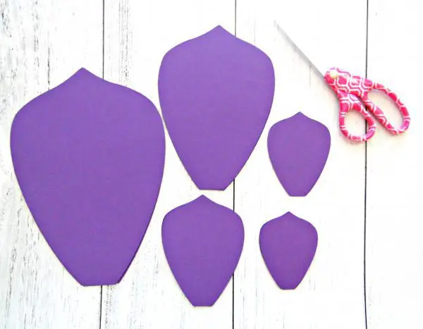 Five purple paper petals of varying sizes lay on a white wood table next to scissors. You can use scissors or a Cricut cutting machine to make these giant paper flowers.