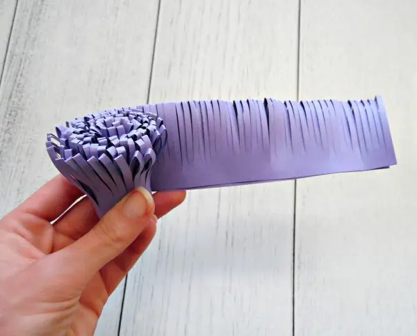Over a white distressed wooden desk, Abbi Kirsten's hand hold a partially rolled and fringed purple piece of paper. This will act as the stamen in a giant paper flower.