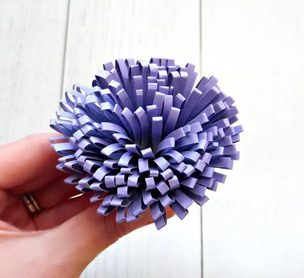 A look at a finished light purple paper pompom held in Abbi Kirsten's hand. The paper stamen pompom will be used when making a giant paper flower from a template.