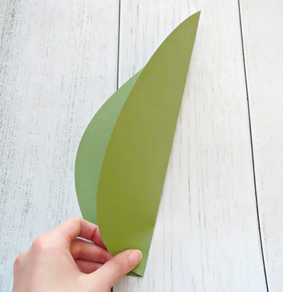 Abbi Kirsten's hand is visible as she folds a large green piece of paper, shaped like a giant leaf, in half. You can add greenery to your giant paper flower.