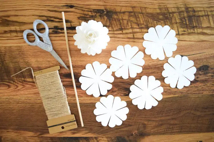 Six pomander paper flower templates cut out, and one completed pomander flower, a wooden dowel, scissors, and twine set out on a wooden table.