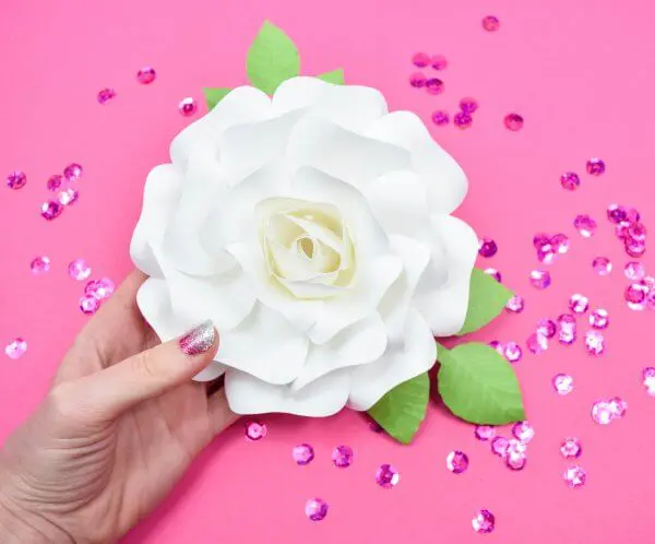 A hand holding a white DIY paper rose embellished with green leaves over a pink table sprinkled with hot pink sequins.