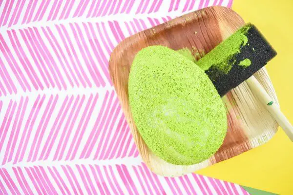 A foam brush sits next to a foam egg painted neon green, on top of a faux wooden plate.