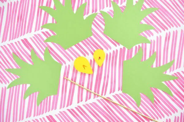 Four green paper pineapple stems, two yellow paper pineapple scales and a bamboo skewer lay on a background of fun pink slashes.