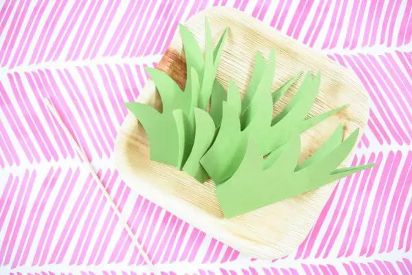 Paper pineapple stems folded in half lay in a square paper faux-wooden craft plate.