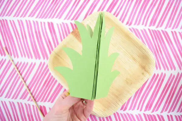 Above a background of pink slashes on white paper, Abbi Kirsten's hand holds a pineapple stem made of four pieces of green paper. After assembly the green paper will make a three-dimensional pineapple crown.