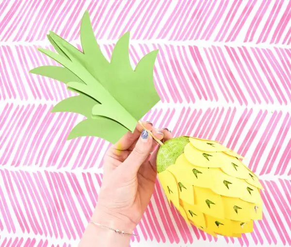 Abbi Kirsten pushes the skewer end of the pineapple stem into the top of a green foam egg covered in paper pineapple scales.