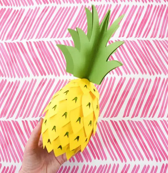 A paper pineapple party decoration craft is held by Abbi Kirsten's hand over a pink and white festive background.