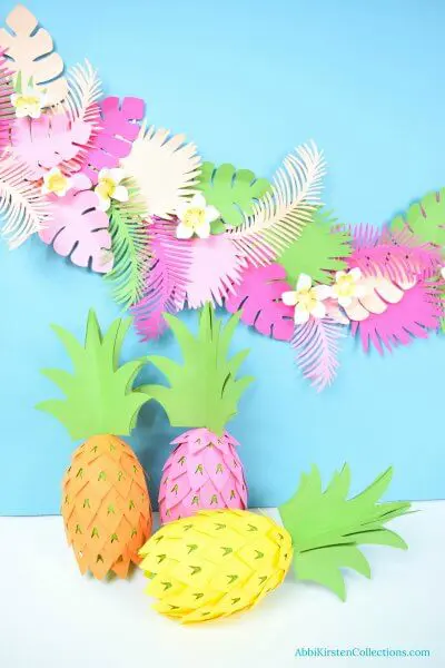Three DIY pineapple party decorations sit on a white surface against a light blue wall. Behind the pineapples is a paper banner of tropical luau party flowers. "AbbiKirstenCollections.com" is typed in the bottom right corner.