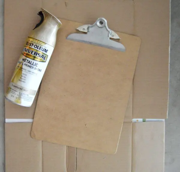 A can of Rustoleum metallic spray lays next to a clipboard on top of cardboard, ready to be painted.