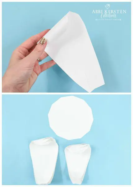 Abbi Kirsten's hand curl the top of the large paper petal inward. Stacks of these curled petals along with a 12 sided base lay on the blue paper tabletop.