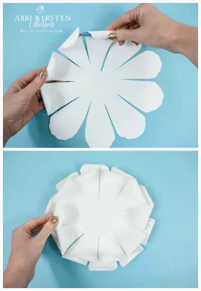 Abbi Kirsten's hands curl the petals of the white petals inward to make the paper flower more realistic. A second image shows the inner curled petals stacked on top of each other.