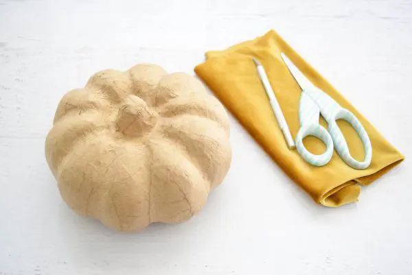 A craft pumpkin, a length of folded gold velvet fabric, scissors, and an Exacto knife are laid out on a table. These supplies are used to make no-sew fabric pumpkins.