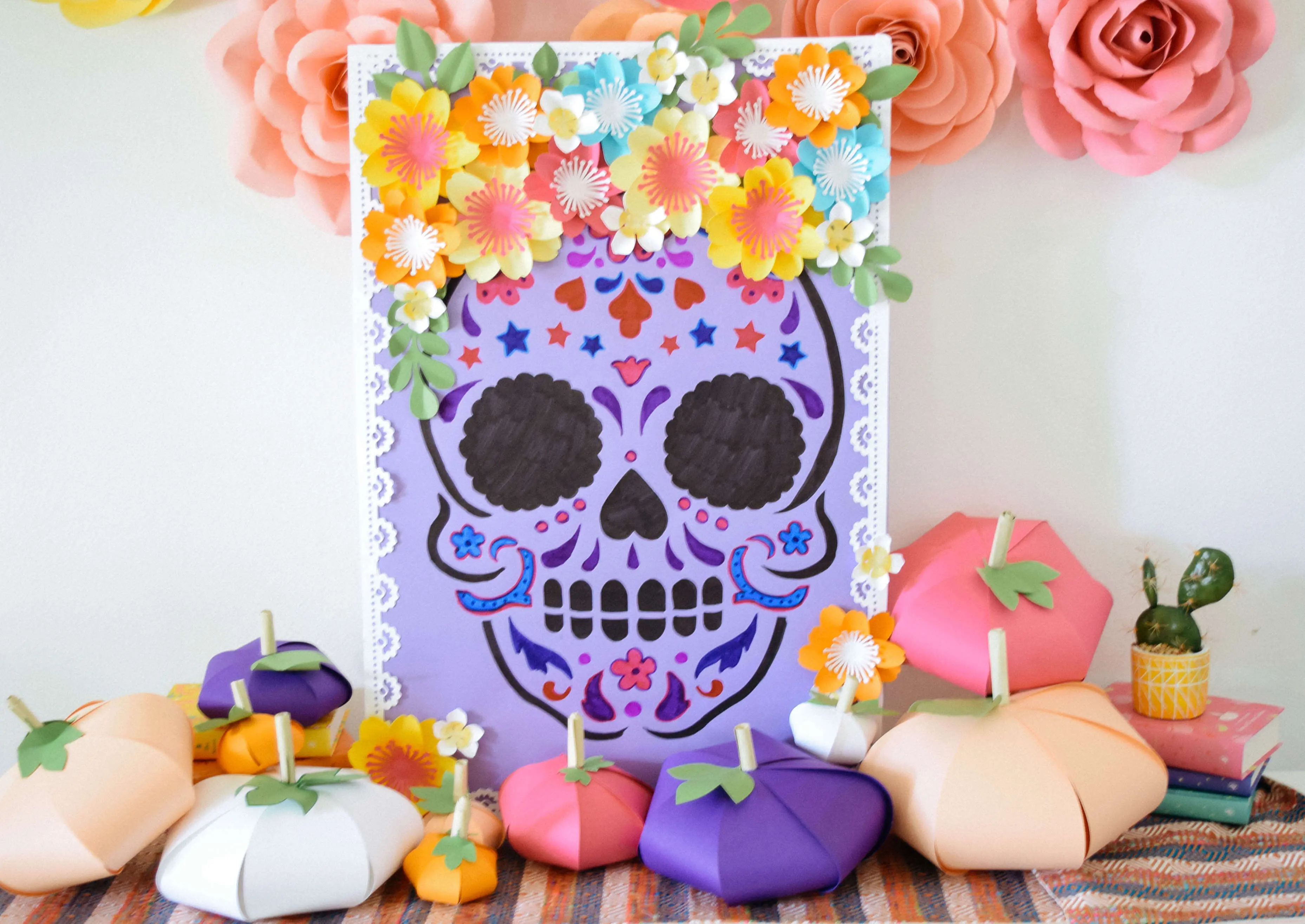 A Day of the Dead sugar skull paper craft is displayed on a Dia de Los Muertos offerings table along with colorful paper pumpkins and giant paper flowers.