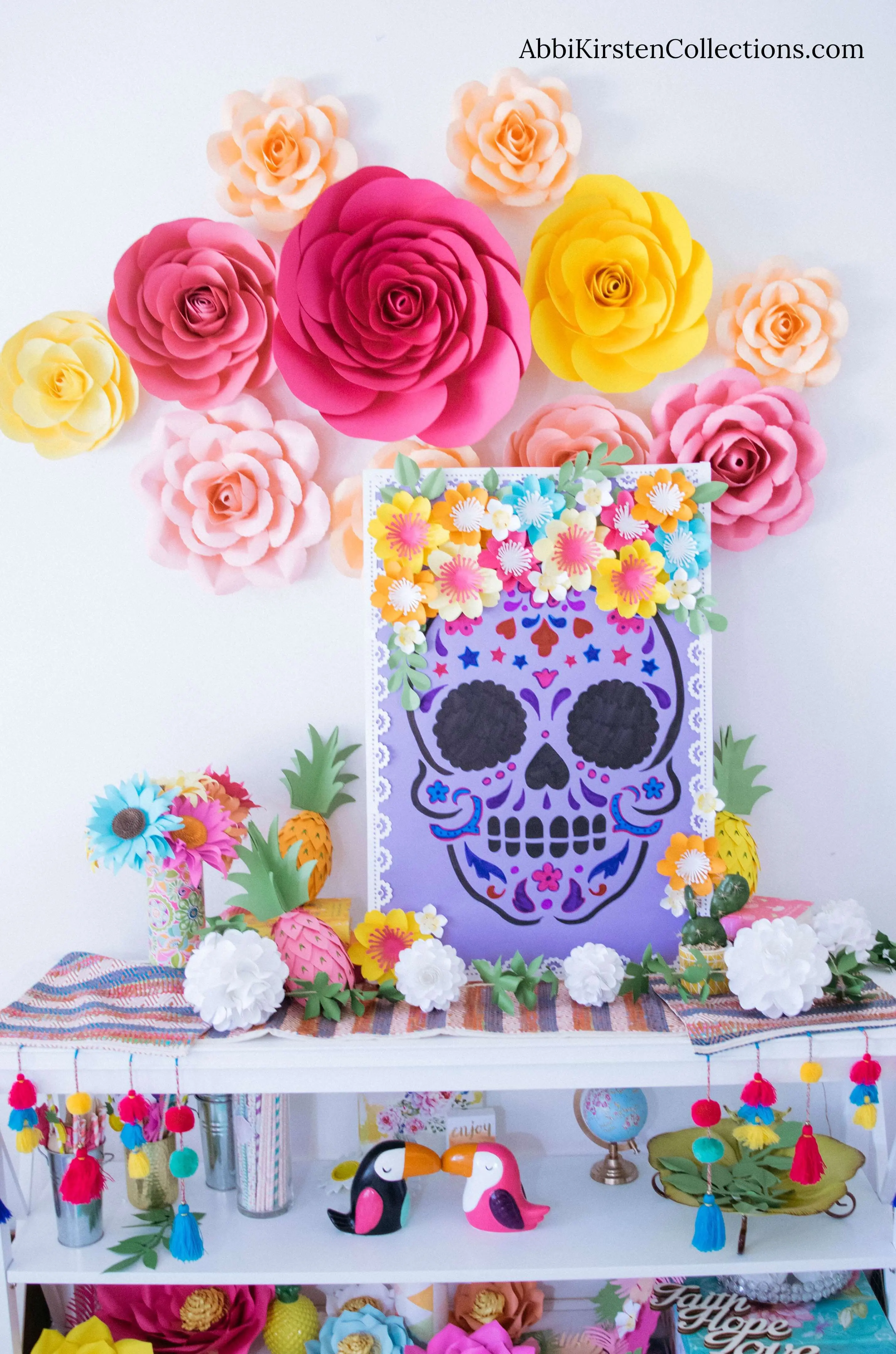 The completed Day of the Dead sugar skull craft is displayed on a table, surrounded by colorful decorations – paper flowers of all shapes and sizes, festive Dia de los Muertos decorations, and giant paper flowers hanging on the walls.