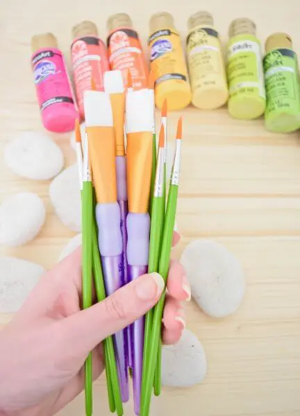 A hand holding a group of paintbrushes. There is paint and rocks in the background. 