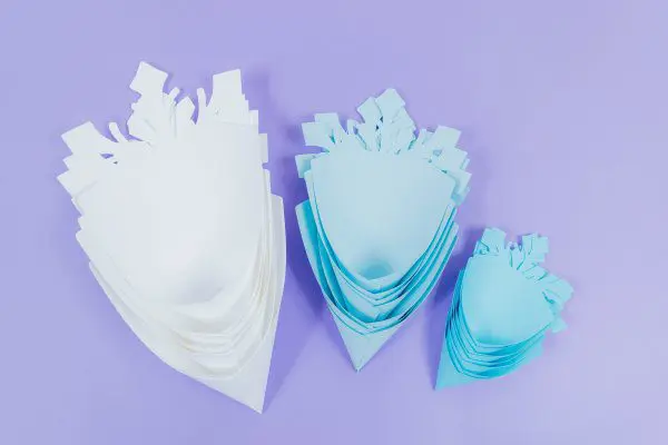 All of the various paper snowflake petals sit together, stacked, after being folded into a cone shape. There are three piles - larger light blue petals, sky blue medium sized petals, and robin's egg blue small petals.