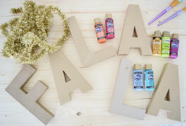 A wooden table full of supplies needed to make DIY decorative tinsel Christmas letters. Supplies include gold tinsel in the corner, above the carboard letters spelling out "Fa La La." Craft paint bottles and brushes round out the supply list.