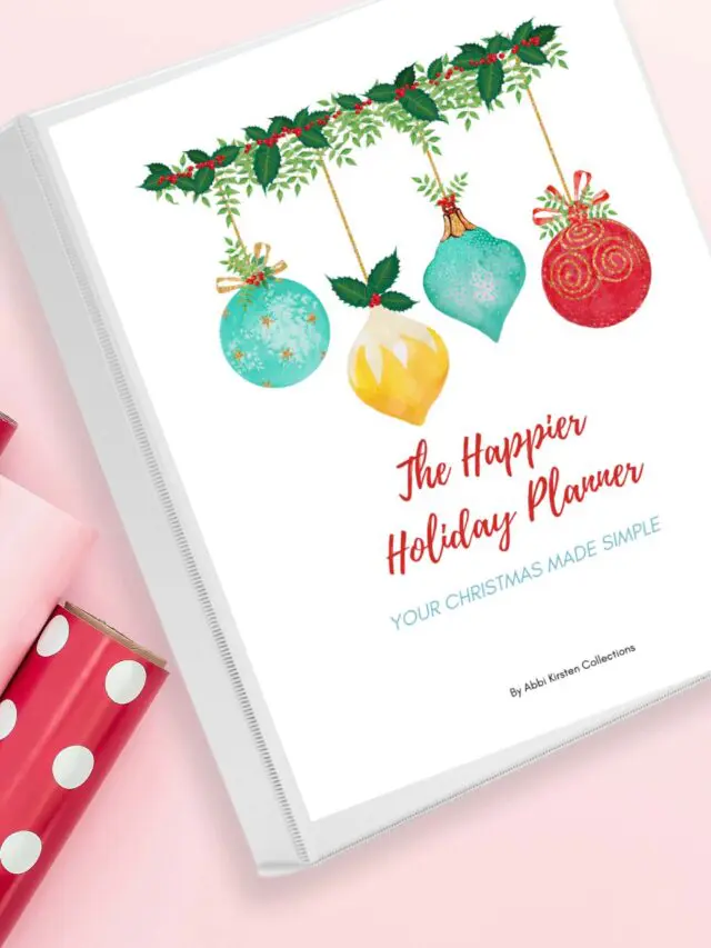 Flat lay photo of The Happier Holiday Planner on a pink surface beside three gift wrapping paper