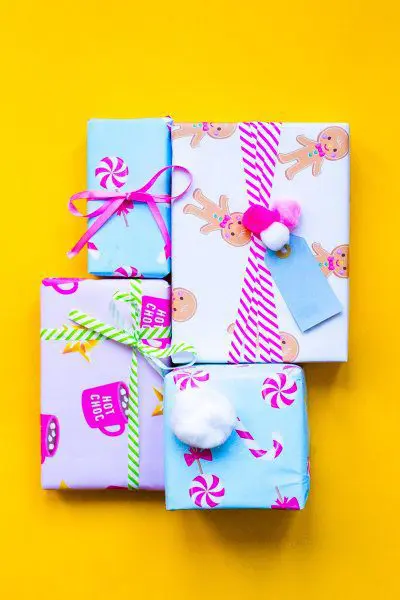 Four gift boxes are wrapped in custom-printed hot chocolate, gingerbread cartoon, and candy-printed wrapping paper with festive striped ribbon, displayed on a bright yellow background.