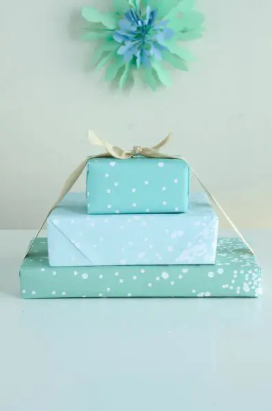 Three blue presents, tied with a yellow ribbon, are sitting on a blue tabletop in front of a blue-green wall. The printable paper is Tiffany-blue with dots of white snow.