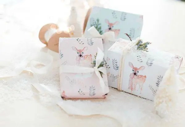 A pile of presents rests on white blankets. These presents are wrapped in pale blue and white wrapping paper with illustrations of a delicate pastel deer, and wrapped with white ribbon.