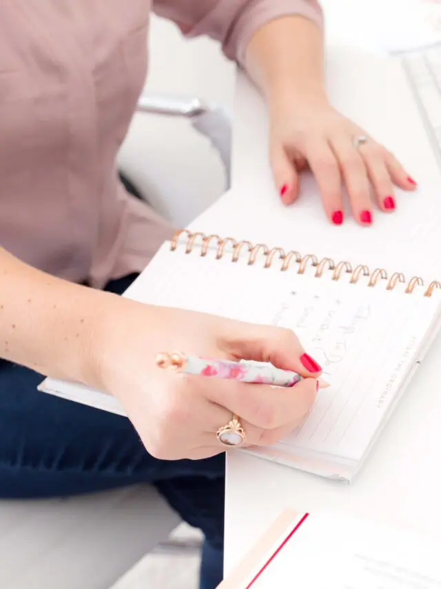A woman wearing a button down shirt and jeans while sitting behind a white desk while writing something on a planner