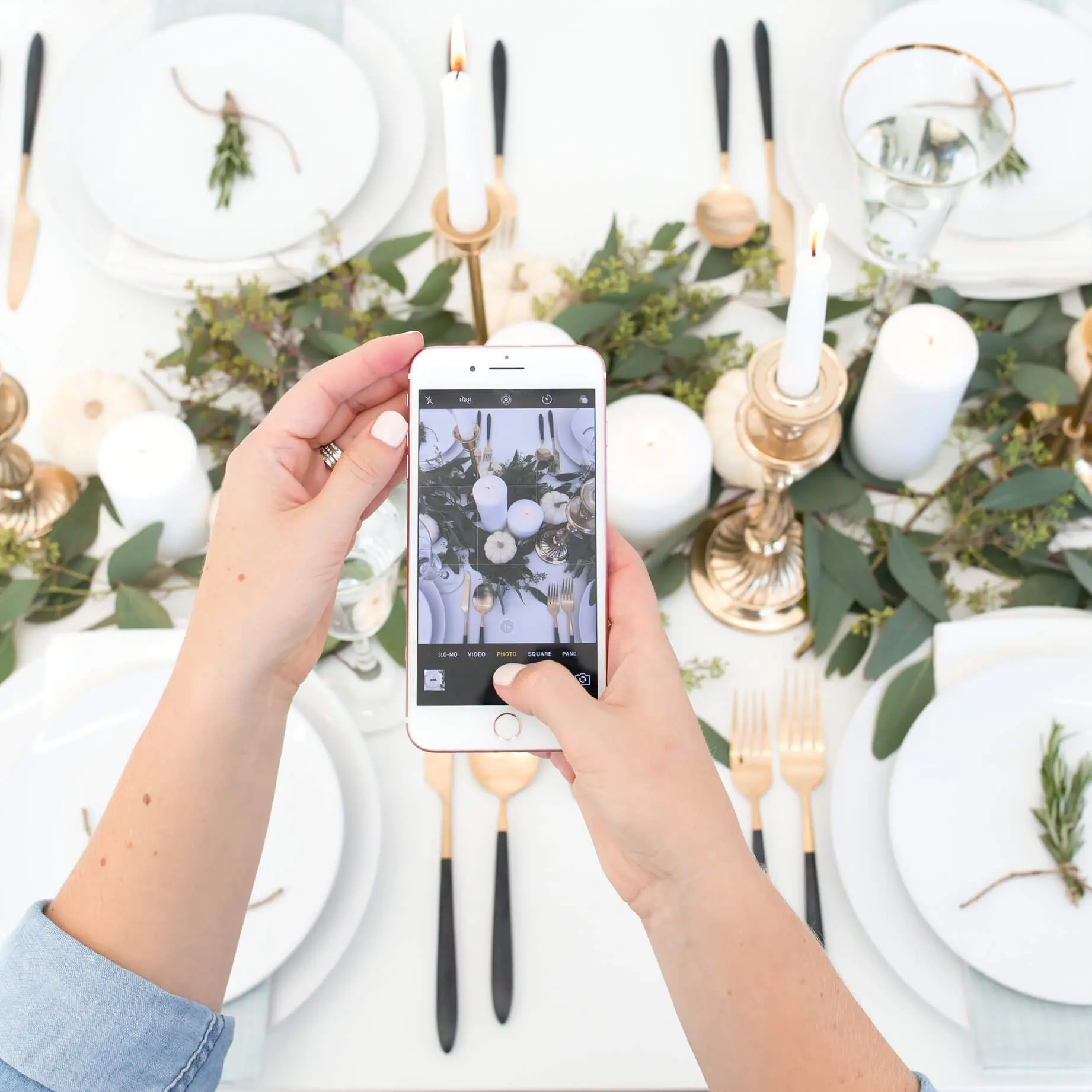 A woman uses her smartphone to take a picture of a beautifully curated holiday tablescape. White dinner plates, gold and black silverware, and gold candlesticks are set up around a greenery centerpiece. 