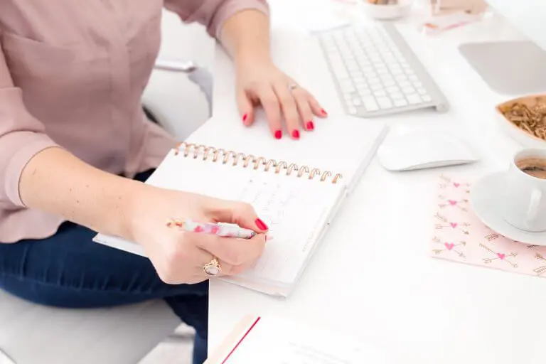A woman wearing a button down shirt and jeans while sitting behind a white desk while writing something on a planner