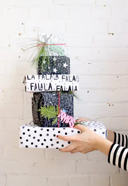 A woman's arms hold a stack of black and white wrapped presents against a white brick wall. Black and white printed gift wrap makes your gift dazzle. Each gift is wrapped with a different patterned paper, from white and black polka dots, stars, and festive “fa la la” print.