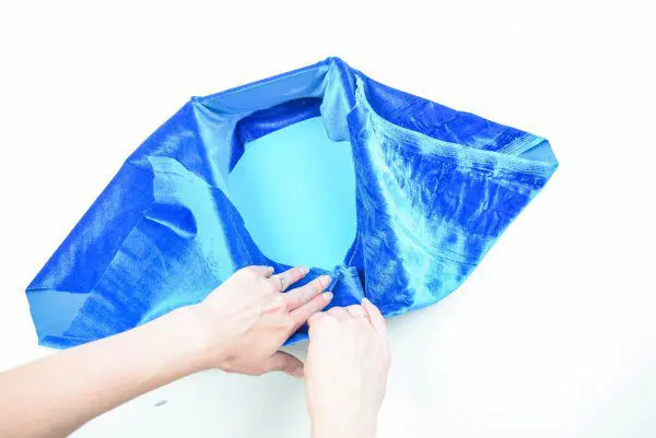 Abbi Kirsten's hands wrap blue velvet around a cardboard box to use as storage. She is tucking the fabric into the corners of the inside of the box.