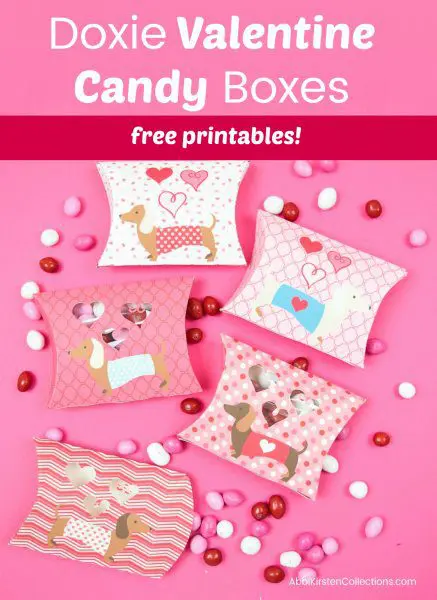 Daschund-themed Valentine's Day pillow boxes lay on a pink background. Holiday-colored candy spills out. The text says, "Doxie Valentine candy boxes free printables!"
